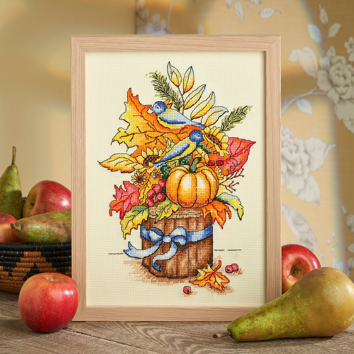 Framed cross-stitched picture of a pumpkin and autumn leaves with fruits on a wooden surface.