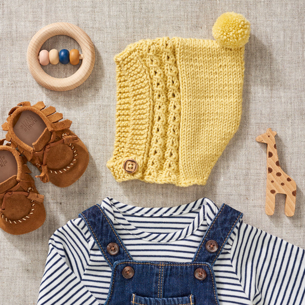 A  yellow pixie bonnet knitting pattern with a pom pom