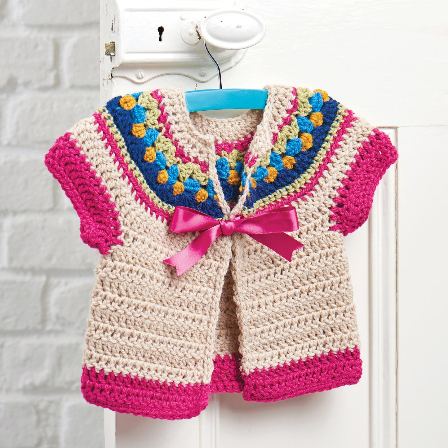 Crocheted baby cardigan with colorful yoke of blue, yellow, and green, pink sleeves and trim, tied with a pink ribbon on a blue hanger.