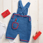 Blue crochet baby overalls with red accents on a wooden surface.