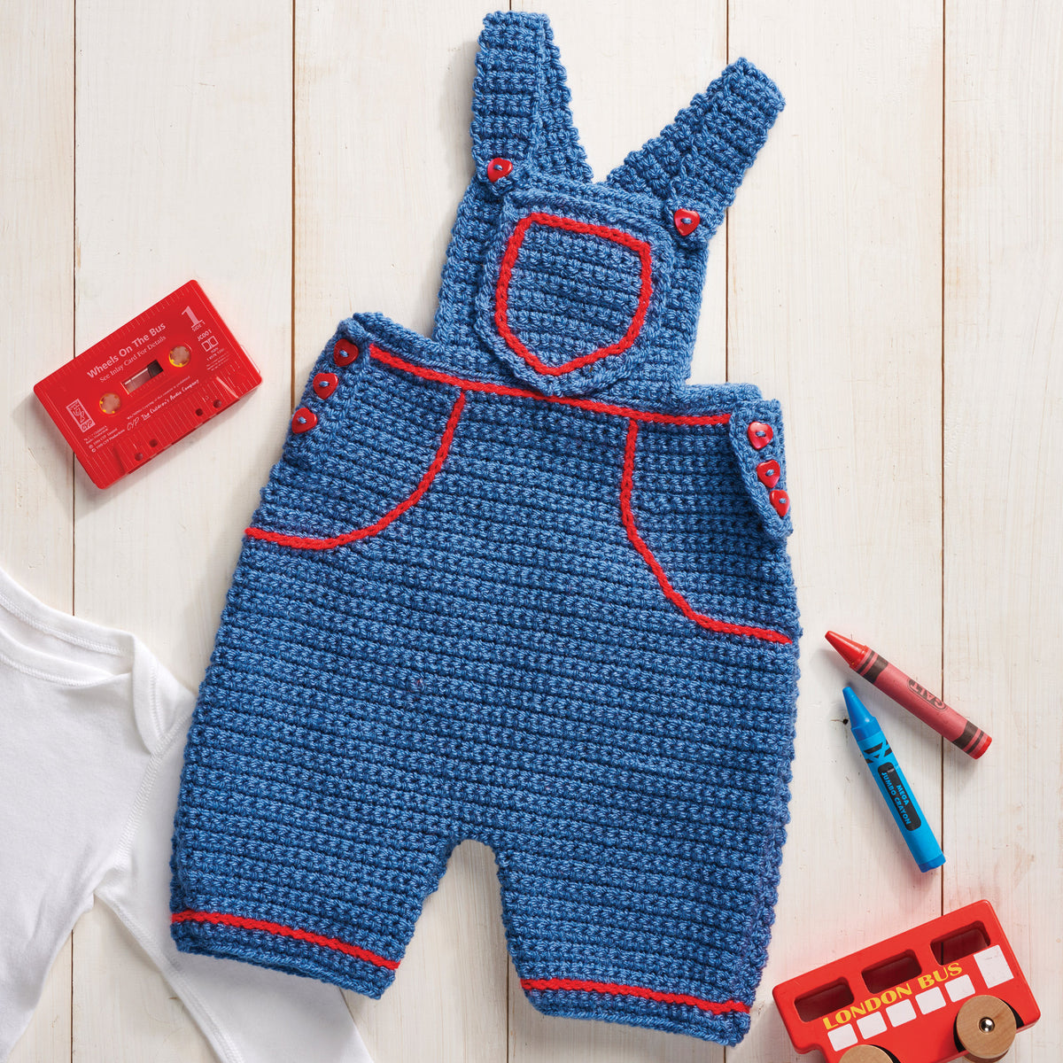 Blue crochet baby overalls with red trim on a wooden surface with toys and a book.