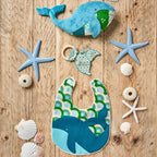 Baby bib, teether, and toy with whale design on a wooden surface with starfish and shells.