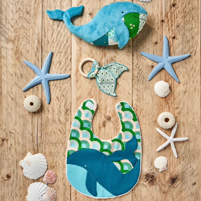Baby bib, teether, and toy with whale design on a wooden surface with starfish and shells.