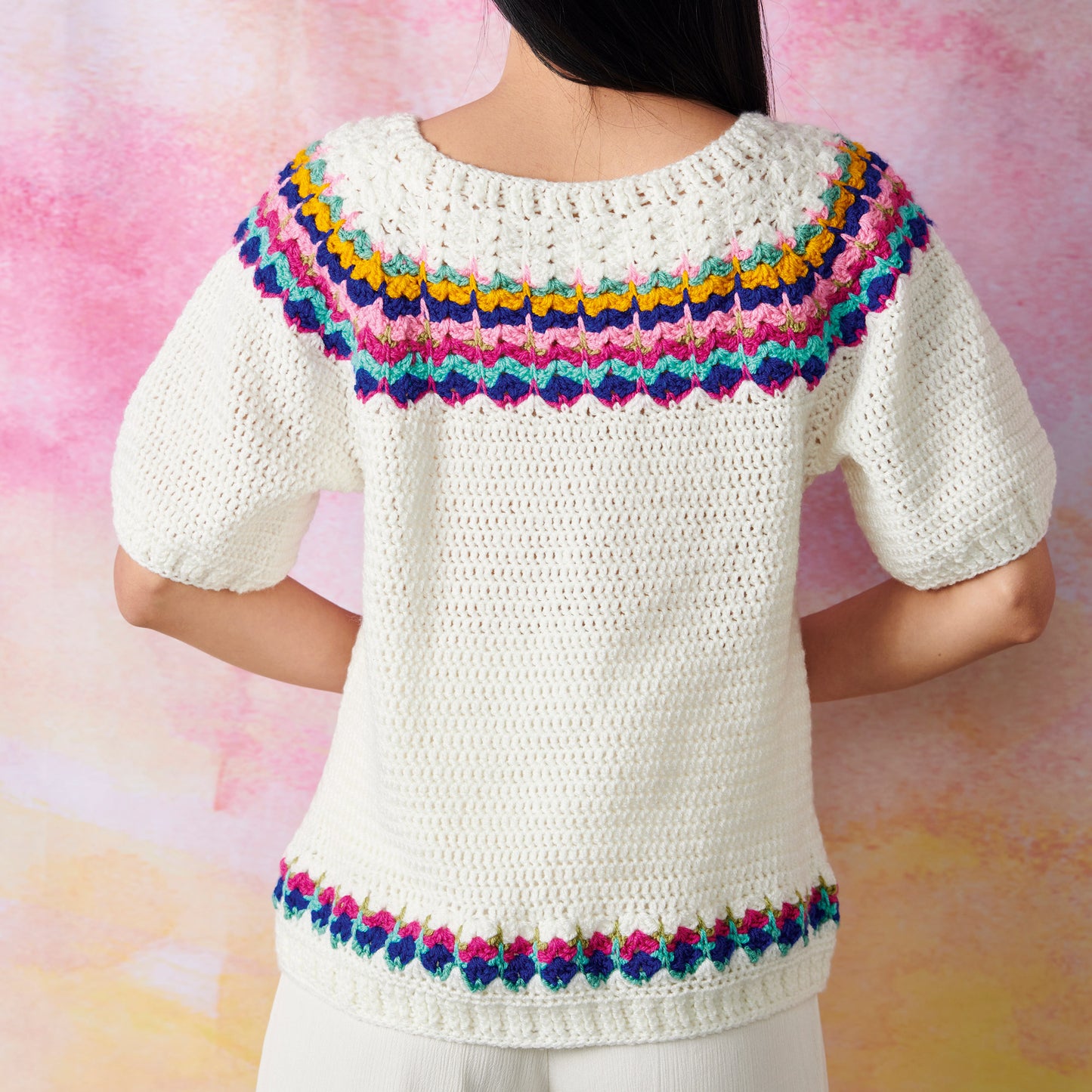 A person wearing a cream crochet sweater with colorful zigzag patterns in blue, pink, and yellow on the yoke and hem, set against a pink background.