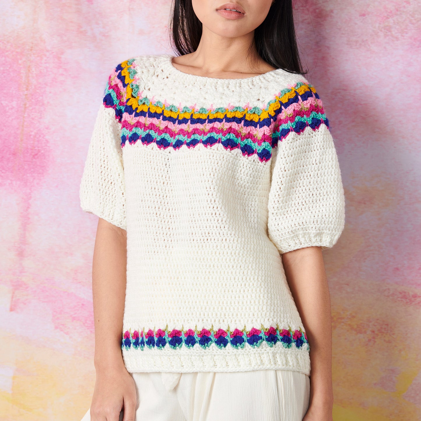 Woman wearing a white knitted sweater with colorful patterns around the neckline and hem. Background is a soft pink and orange gradient wall.