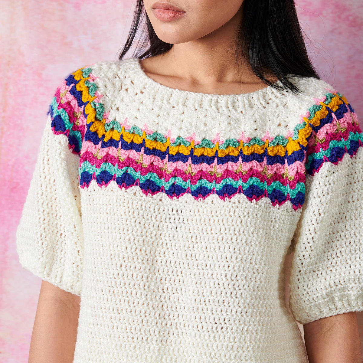A woman wearing a cream-colored crochet sweater with a vibrant, colorful pattern around the neckline. The background is a soft pink gradient, creating a warm, cozy feel.