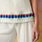 Close-up of a person wearing a cream crocheted top with a colorful floral border in pink and blue. The top is paired with loose, cream trousers.