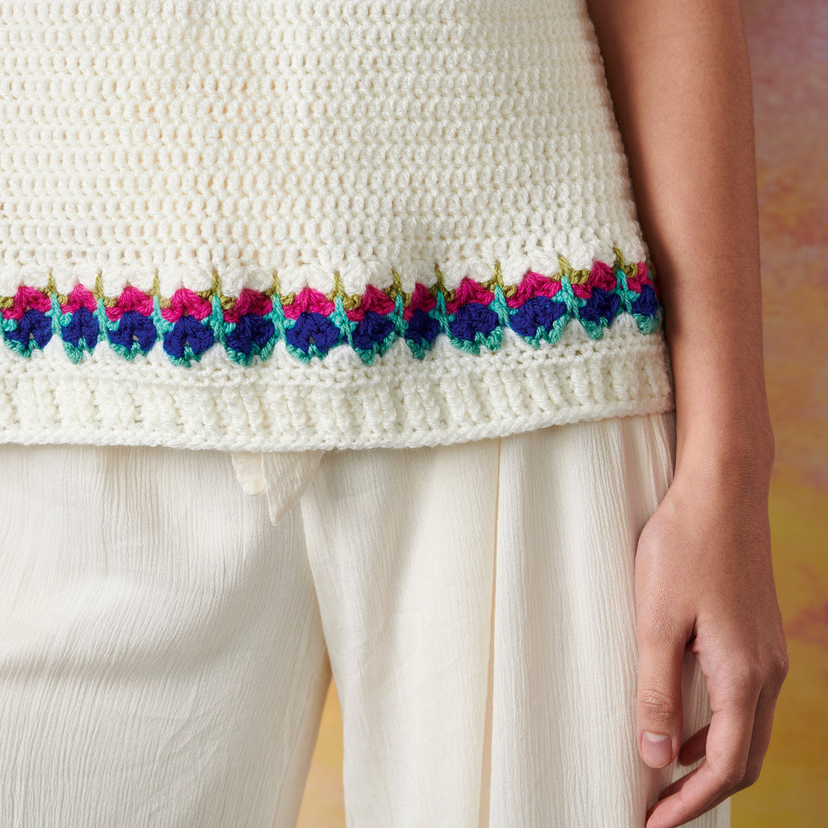 Close-up of a person wearing a cream crocheted top with a colorful floral border in pink and blue. The top is paired with loose, cream trousers.