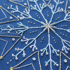 Close-up of an embroidered snowflake design on blue fabric with white and gold thread.