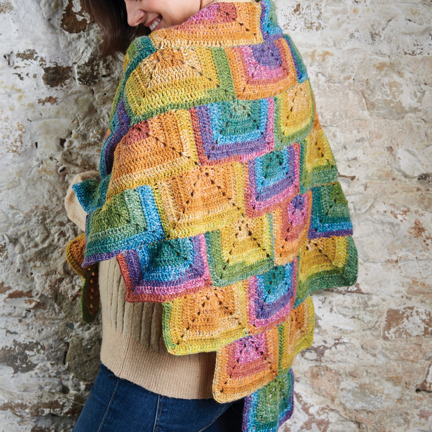 Person wearing a colorful crocheted shawl against a textured wall.