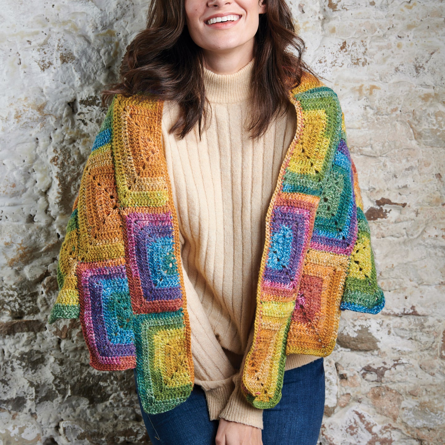 Woman wearing a colorful crocheted cardigan against a stone wall.