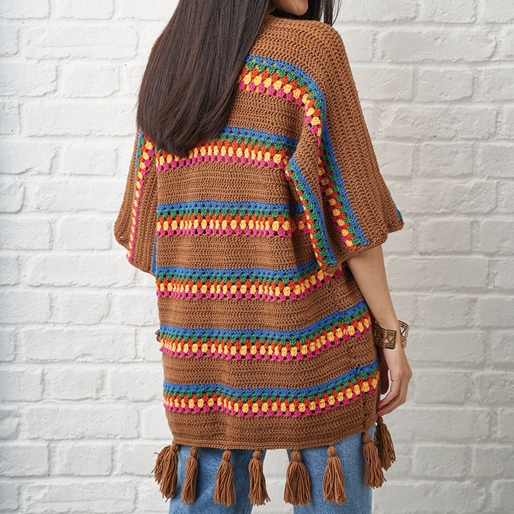 Person wearing a brown crochet poncho with colorful patterns against a white brick wall.