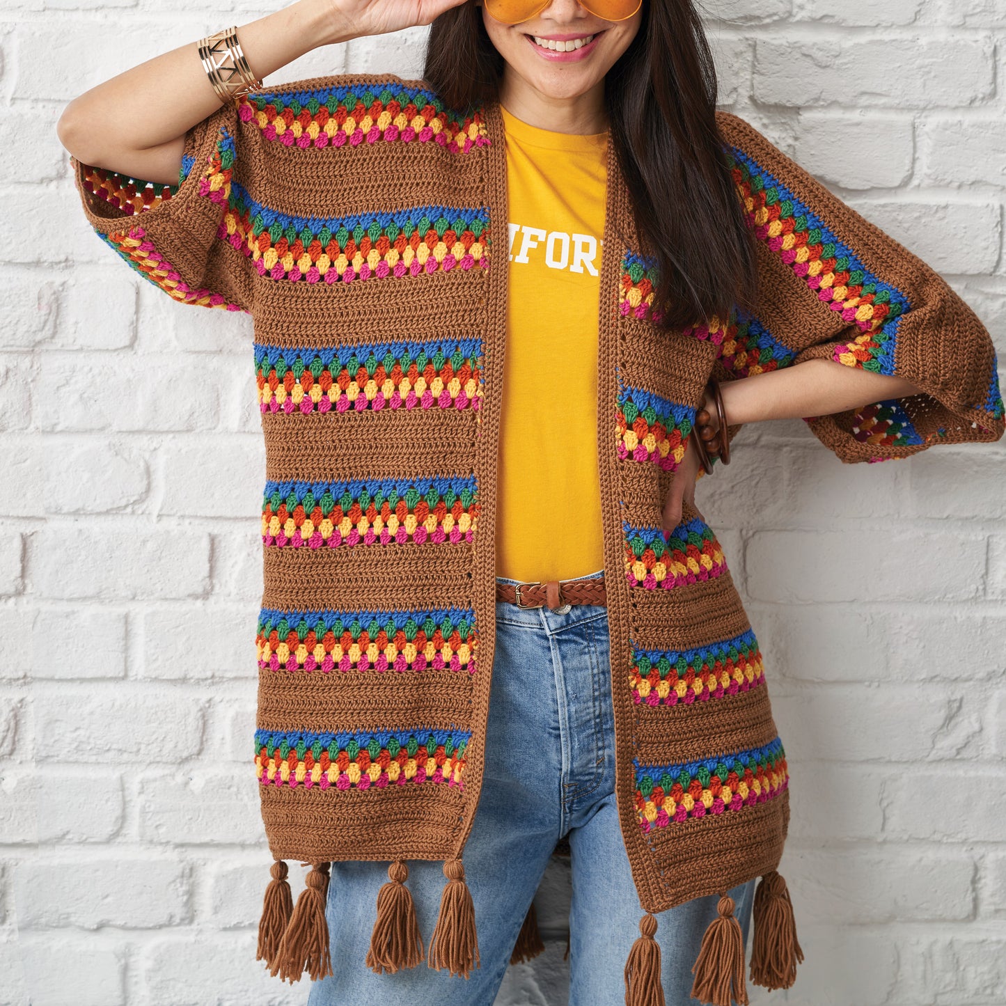 Person wearing a colorful striped cardigan with tassels against a white brick wall.
