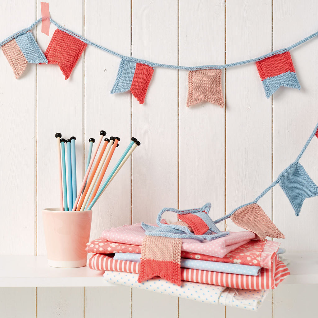 A knitting pattern PDF for a flag bunting design, with knitted flags in red, blue, and white colors displayed on a white wooden background, alongside a collection of knitting needles and yarn.