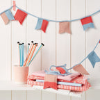 A knitting pattern PDF for a flag bunting design, with knitted flags in red, blue, and white colors displayed on a white wooden background, alongside a collection of knitting needles and yarn.