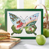 Framed cross-stitched butterfly with fairy design on a table with food items.