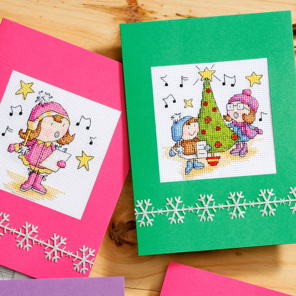 Two cross-stitched cards on a wooden surface, one pink with a girl and stars, the other green with children and a tree.