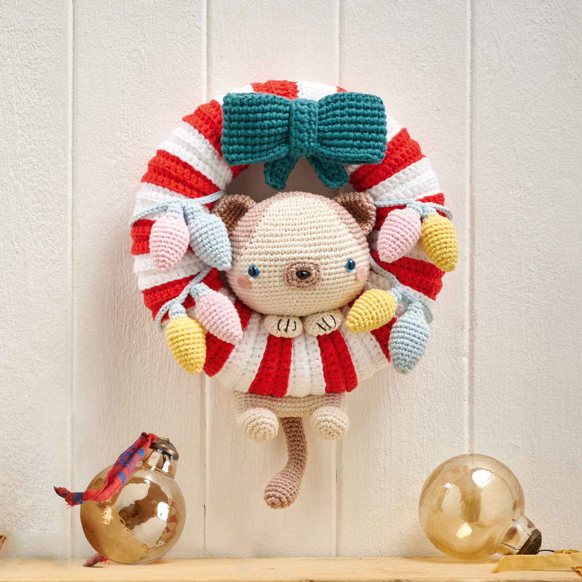 A colorful crochet Christmas wreath featuring a cat figure with a red and white striped pattern, surrounded by various decorative elements like a blue bow and yellow ornaments.