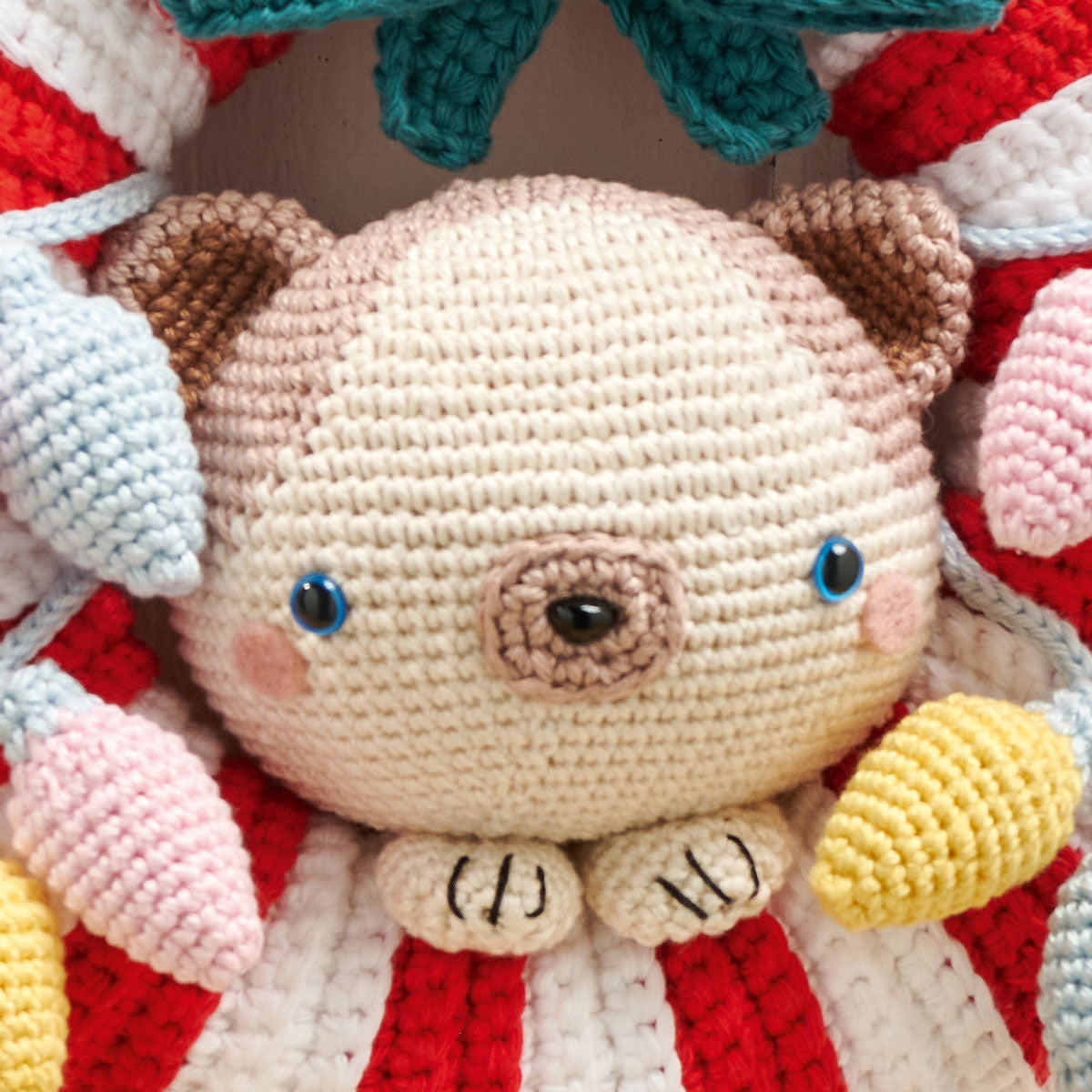 Crocheted cat face with blue eyes and rosy cheeks, surrounded by colorful knitted elements in red, white, pink, and yellow. Playful and cozy atmosphere.