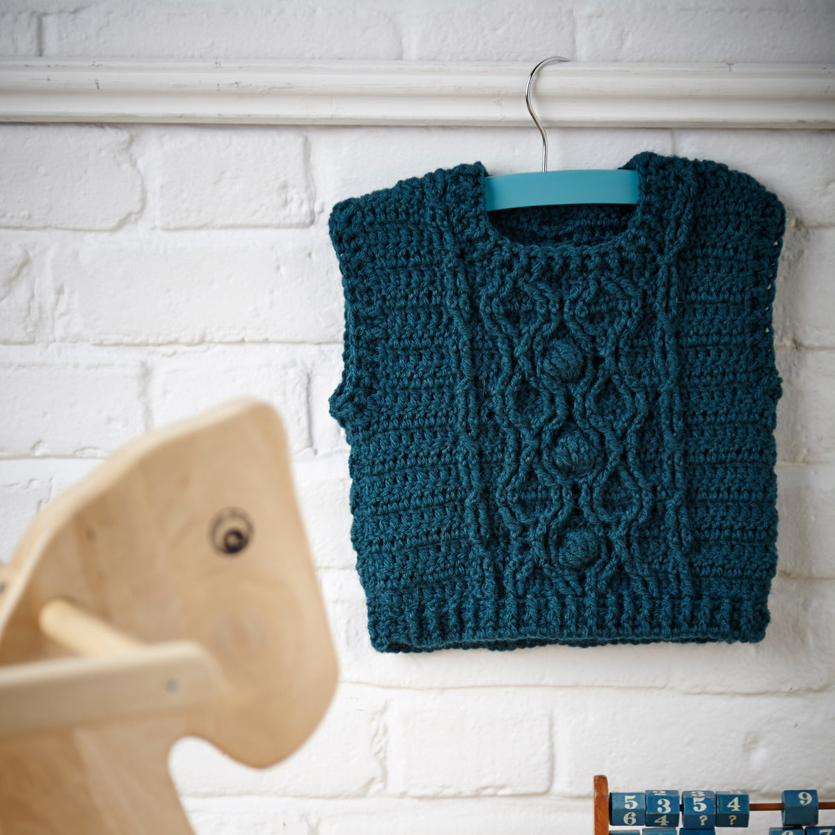 Teal crocheted vest hanging on a white brick wall