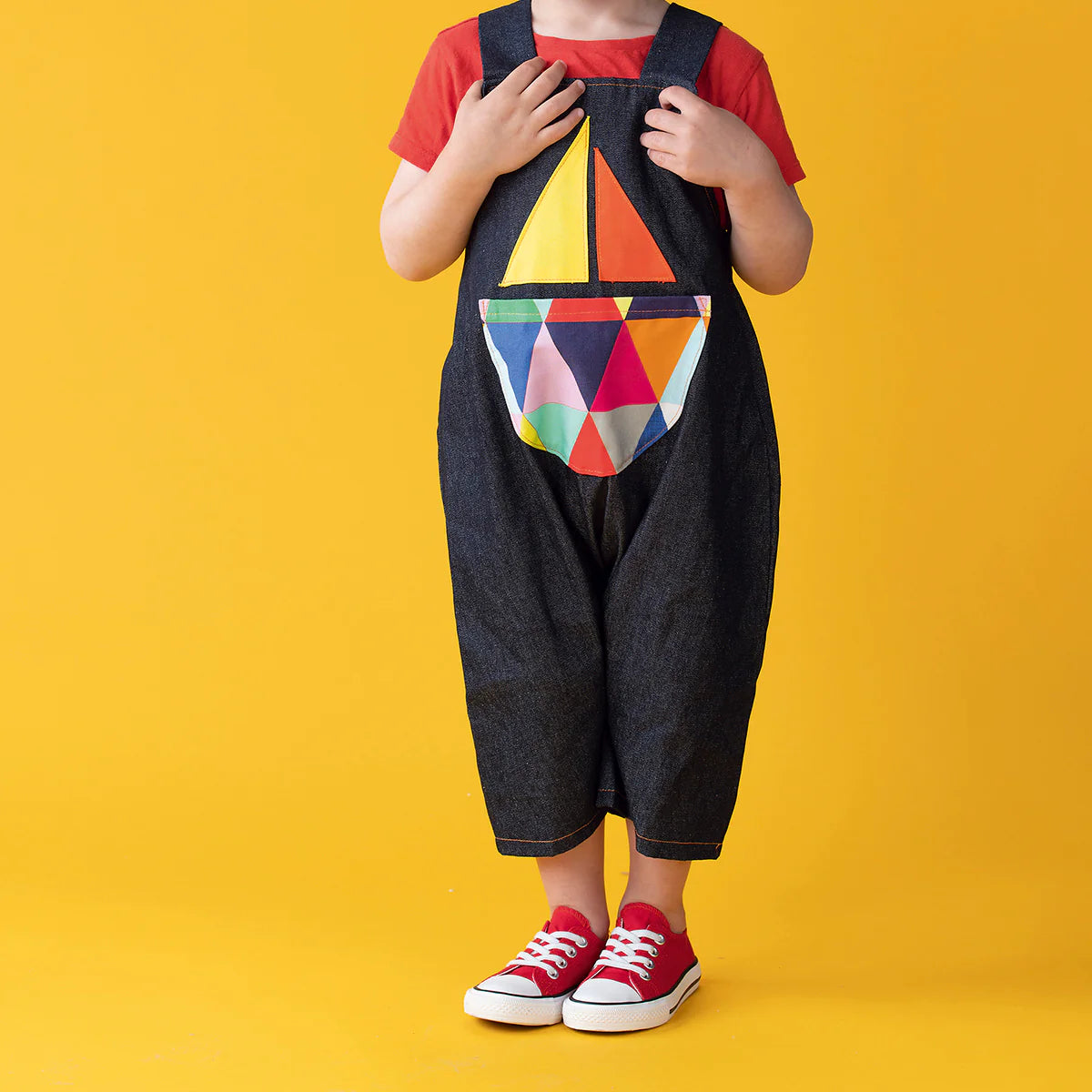 Child wearing a colorful geometric-patterned bib overalls on a yellow background