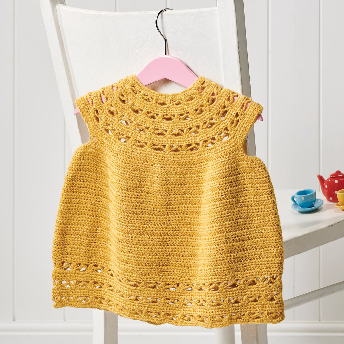 Yellow crocheted dress on a white chair with a pink hanger.