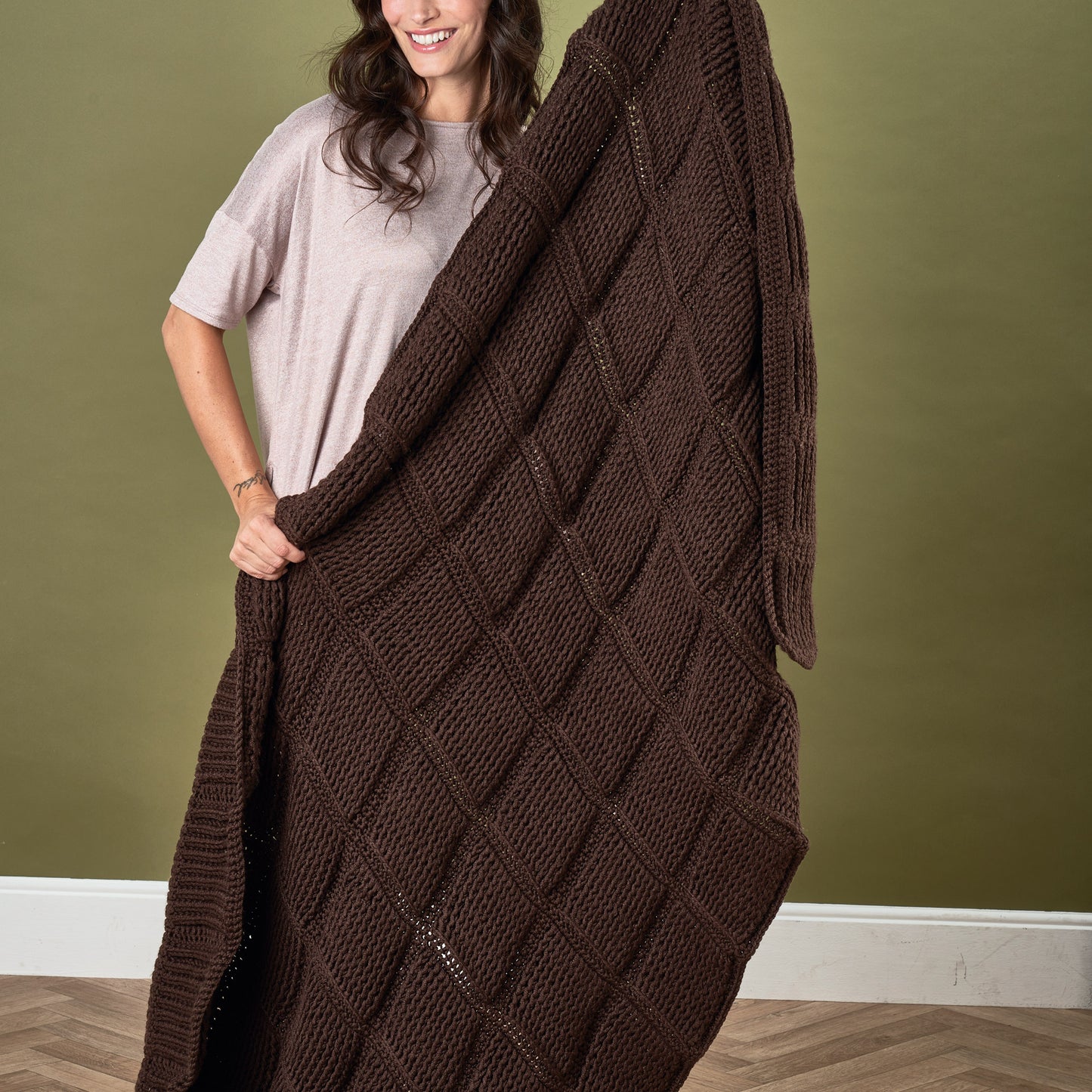 Woman holding a large brown crochet blanket against a green wall.