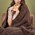 Person wrapped in a brown blanket eating a gold leaf wrapped chocolate bar against a green background