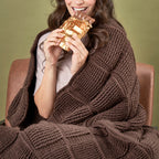 Person wrapped in a brown blanket eating a gold leaf wrapped chocolate bar against a green background