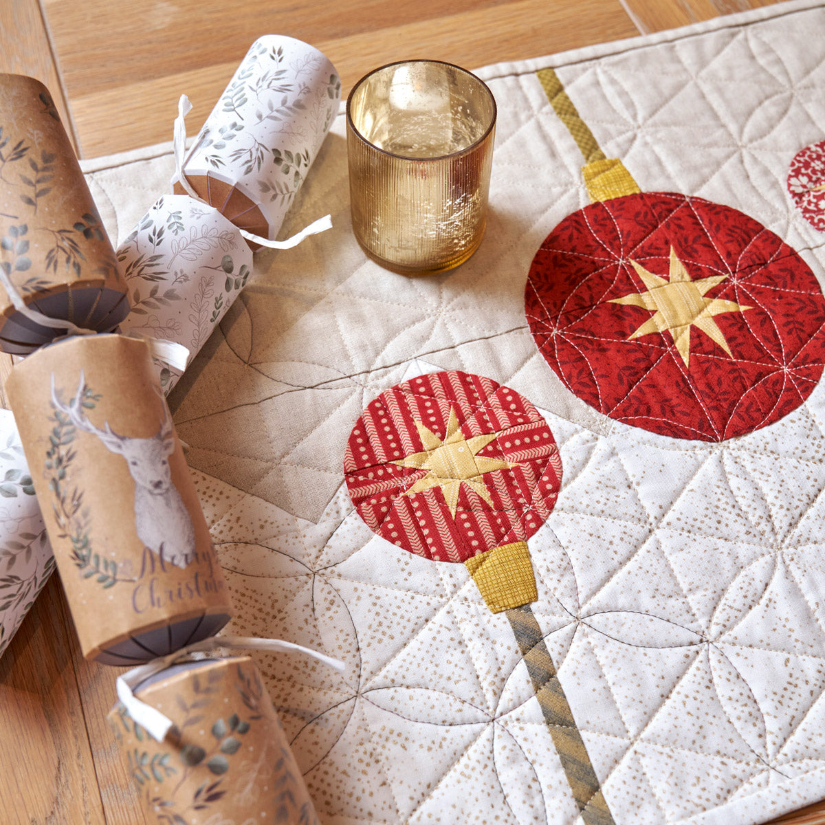 Decorative table setting with quilted placemats, candles, and a glass on a wooden surface.