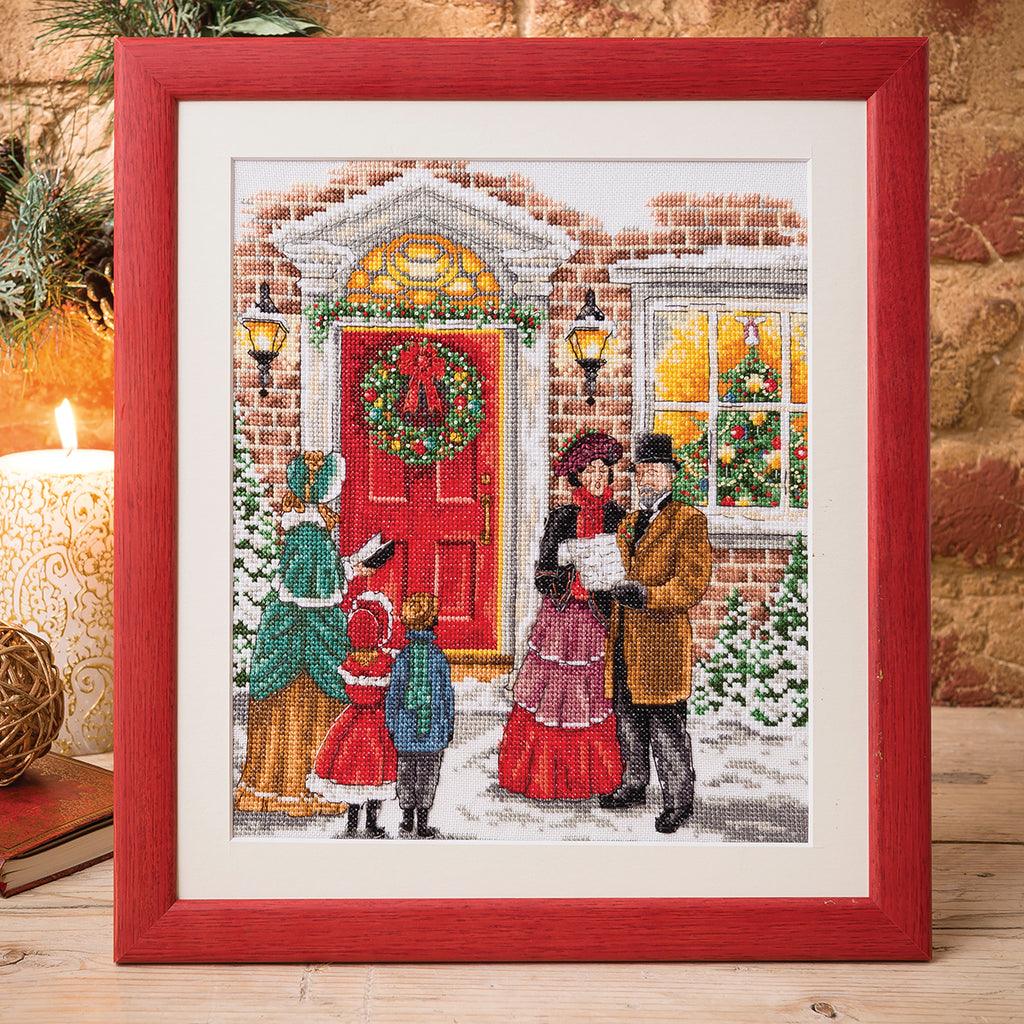 Framed cross-stitched picture of a family in front of a house with a wreath, on a wooden surface.