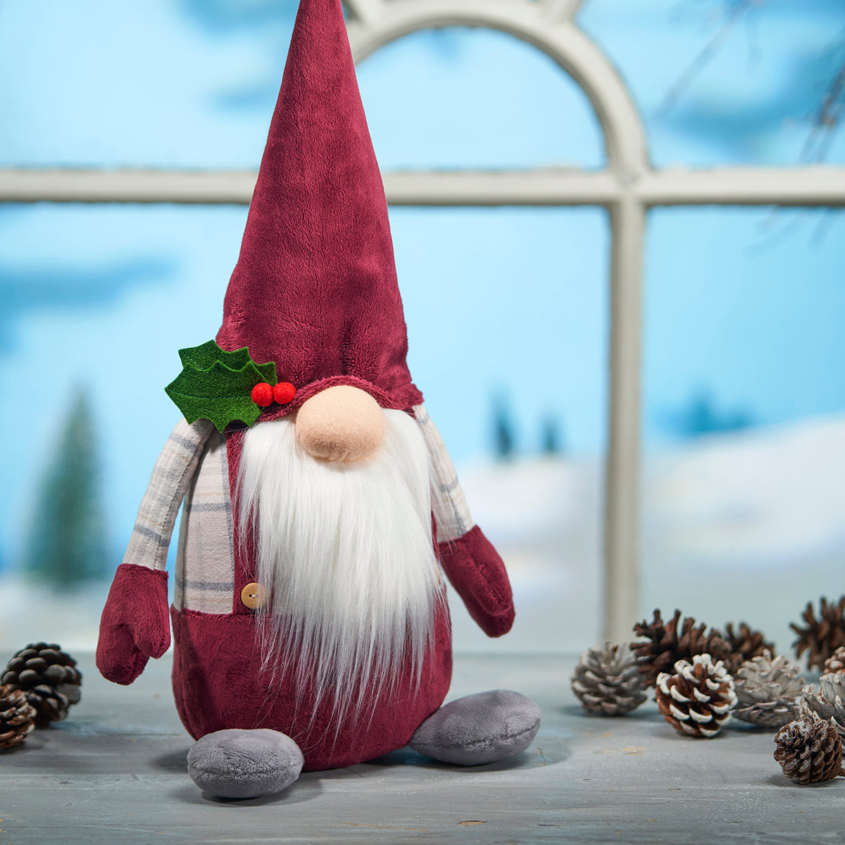 Plush gnome toy with a red hat and white beard, surrounded by pine cones on a wooden surface.