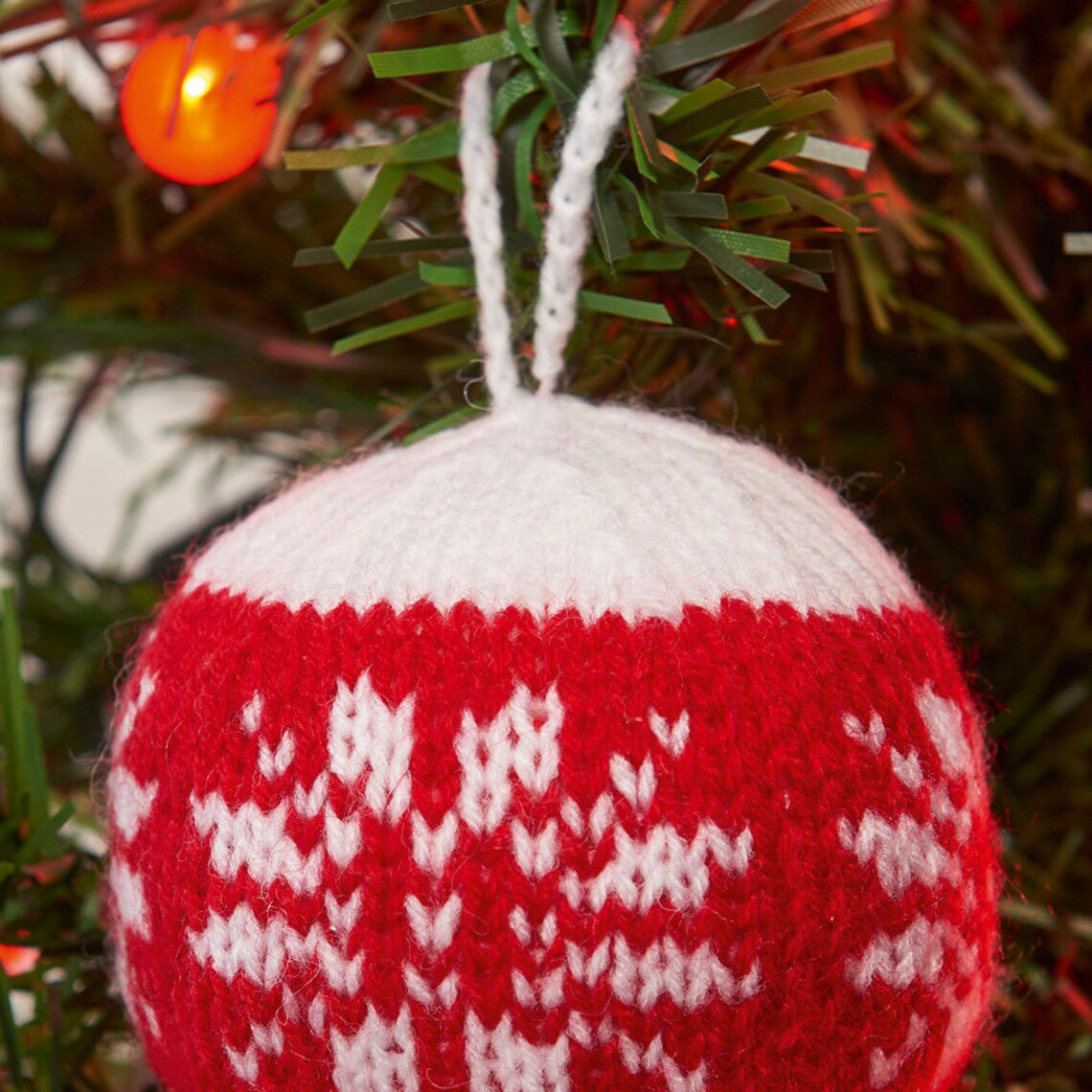 A knitted Christmas bauble in red and white, with a bold, geometric pattern, hanging on a Christmas tree.
