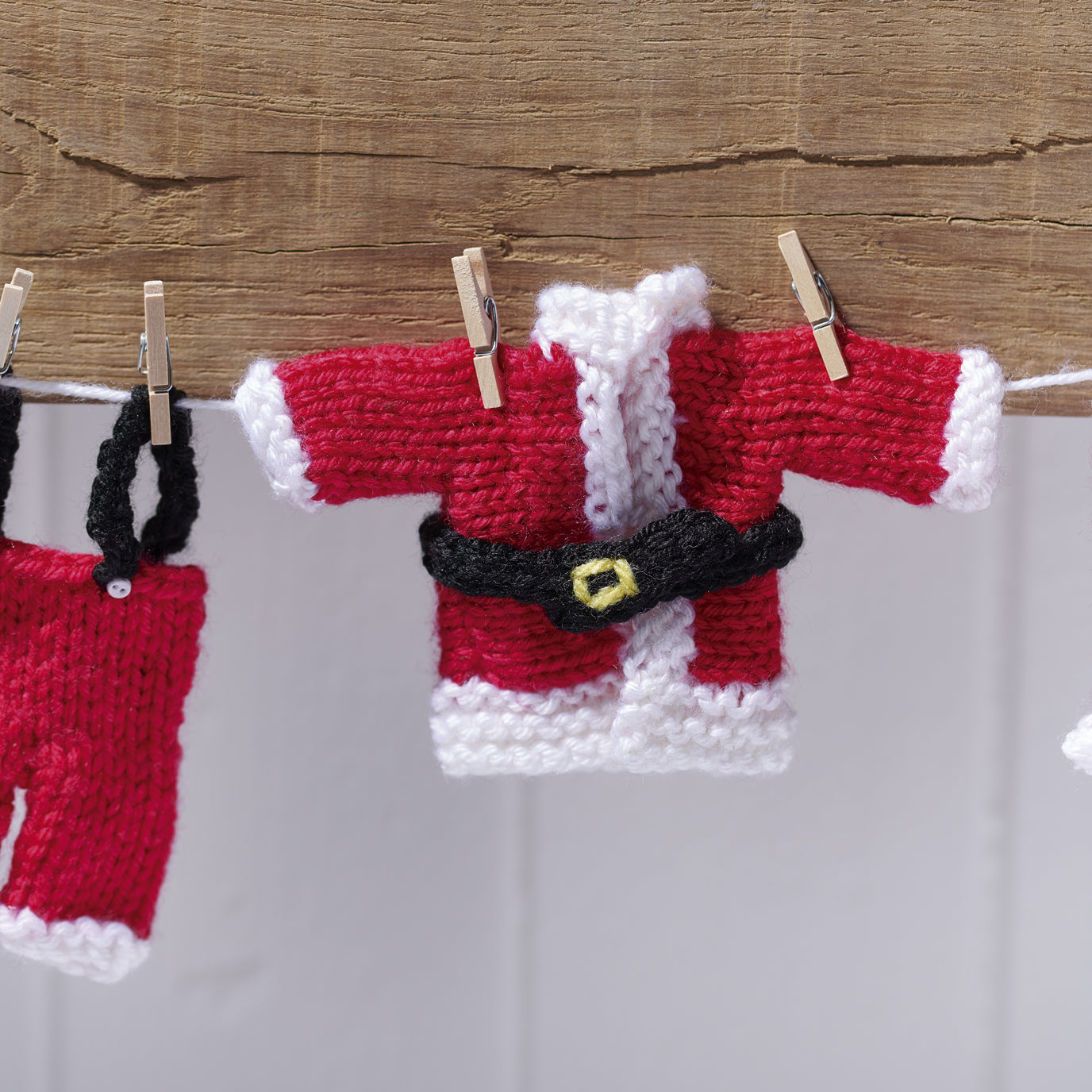 Knitted Santa Claus outfit hanging on a line with wooden pegs against a wooden background.