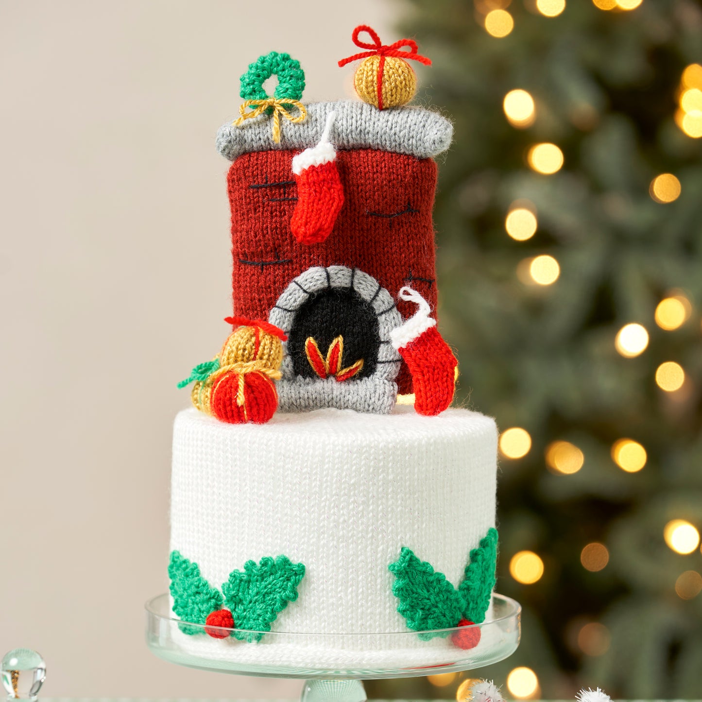 Knitted fireplace cake topper with Santa Claus and holly decorations against a blurred Christmas tree background.