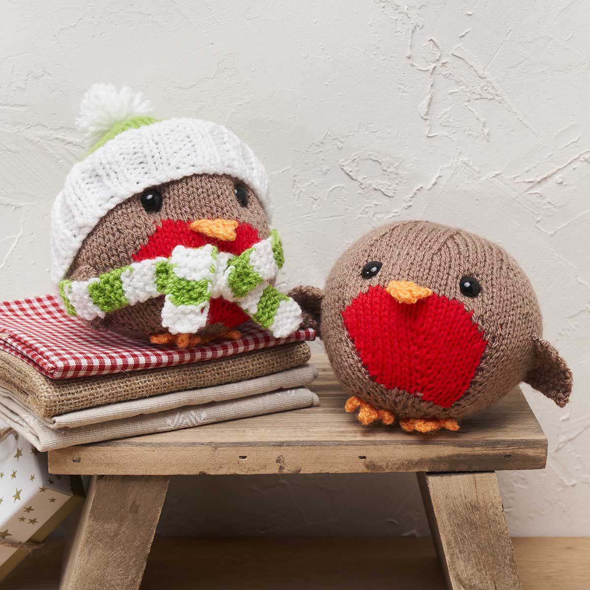 Two knitted robins on a wooden stool against a textured wall.