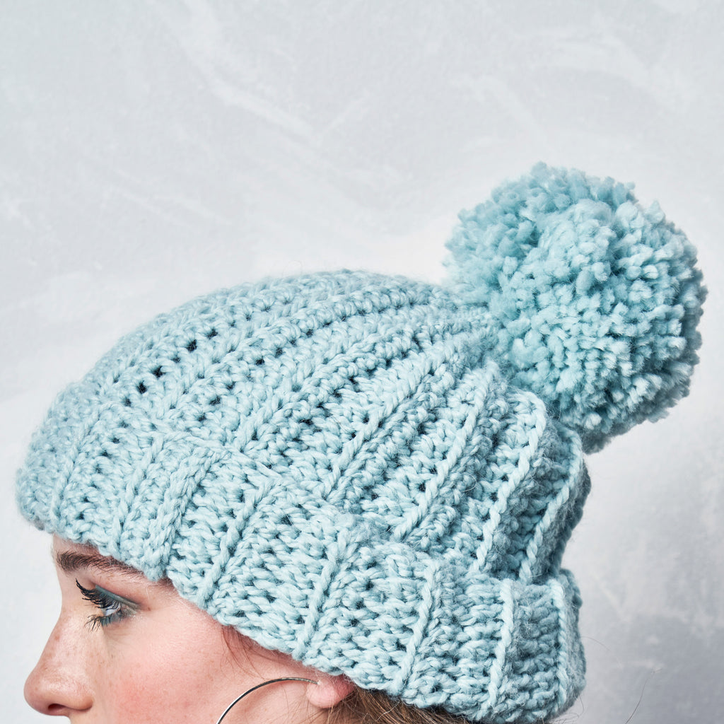 Light blue crochet beanie with a pom-pom on a neutral background