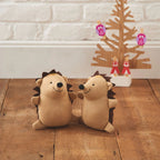 Two plush hedgehogs on a wooden floor with a decorative Christmas tree in the background.