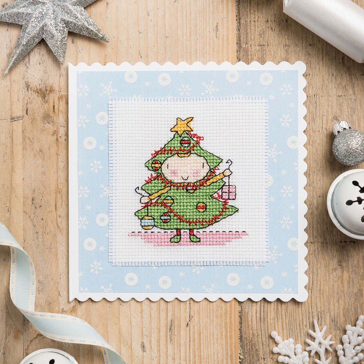 Cross-stitched design of a Christmas tree character on a wooden surface with festive decorations.