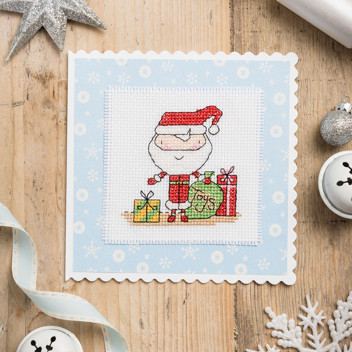 Cross-stitched pattern of Santa Claus with presents on a blue background, surrounded by Christmas decorations.