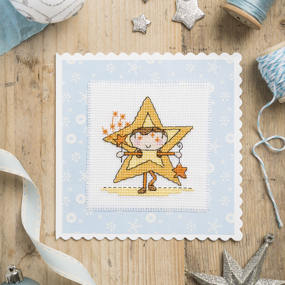 Cross-stitched design of a star-shaped character on a wooden surface with crafting materials.