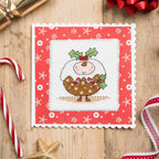 Cross-stitched design of a Christmas pudding on a red card with snowflake pattern, surrounded by festive items on a wooden surface.