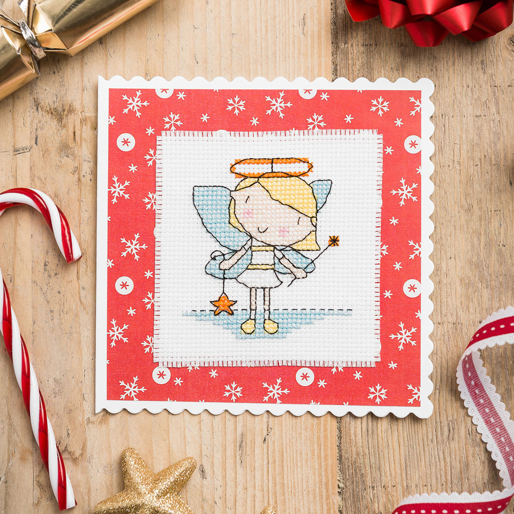 Cross-stitched angel design on a red card with snowflakes, surrounded by Christmas decorations on a wooden surface.