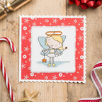 Cross-stitched angel design on a red card with snowflakes, surrounded by Christmas decorations on a wooden surface.