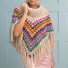 Woman wearing a colorful, crocheted poncho with fringe against a light blue paneled wall. The poncho has a cowl neckline and vibrant geometric patterns.