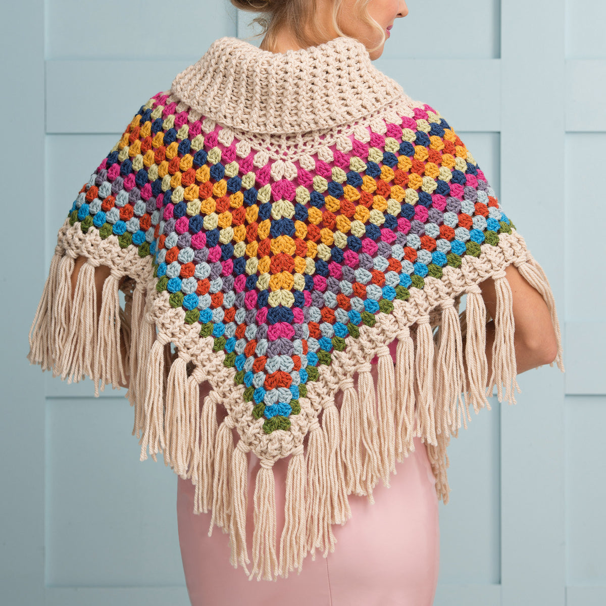 A woman is wearing a colorful crochet shawl with a fringe. The shawl features vibrant diamond patterns in blue, red, yellow, and green, with a beige collar.