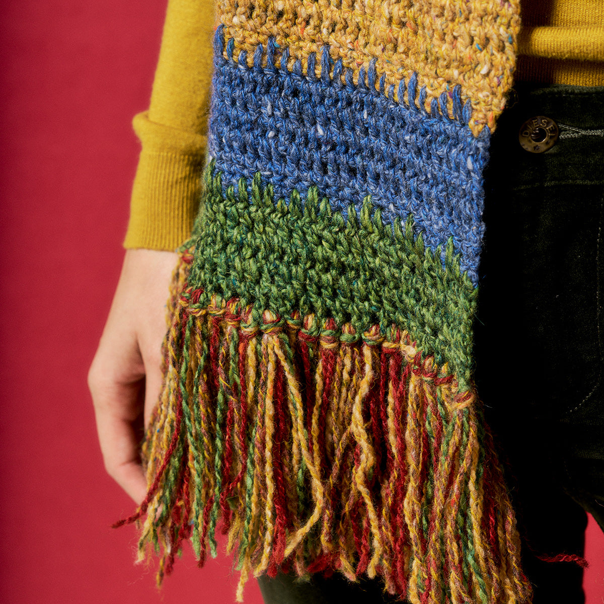 Colorful knitted scarf with fringe held by a person against a red background