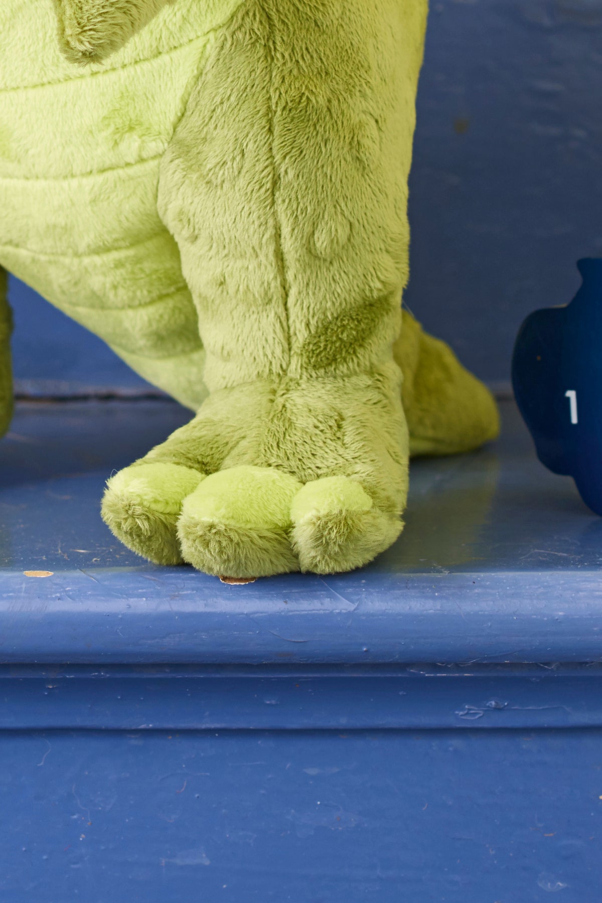 Close-up of a plush green dinosaur's foot with soft, rounded toes, resting on a blue wooden step. The texture is fluffy and the scene is playful.