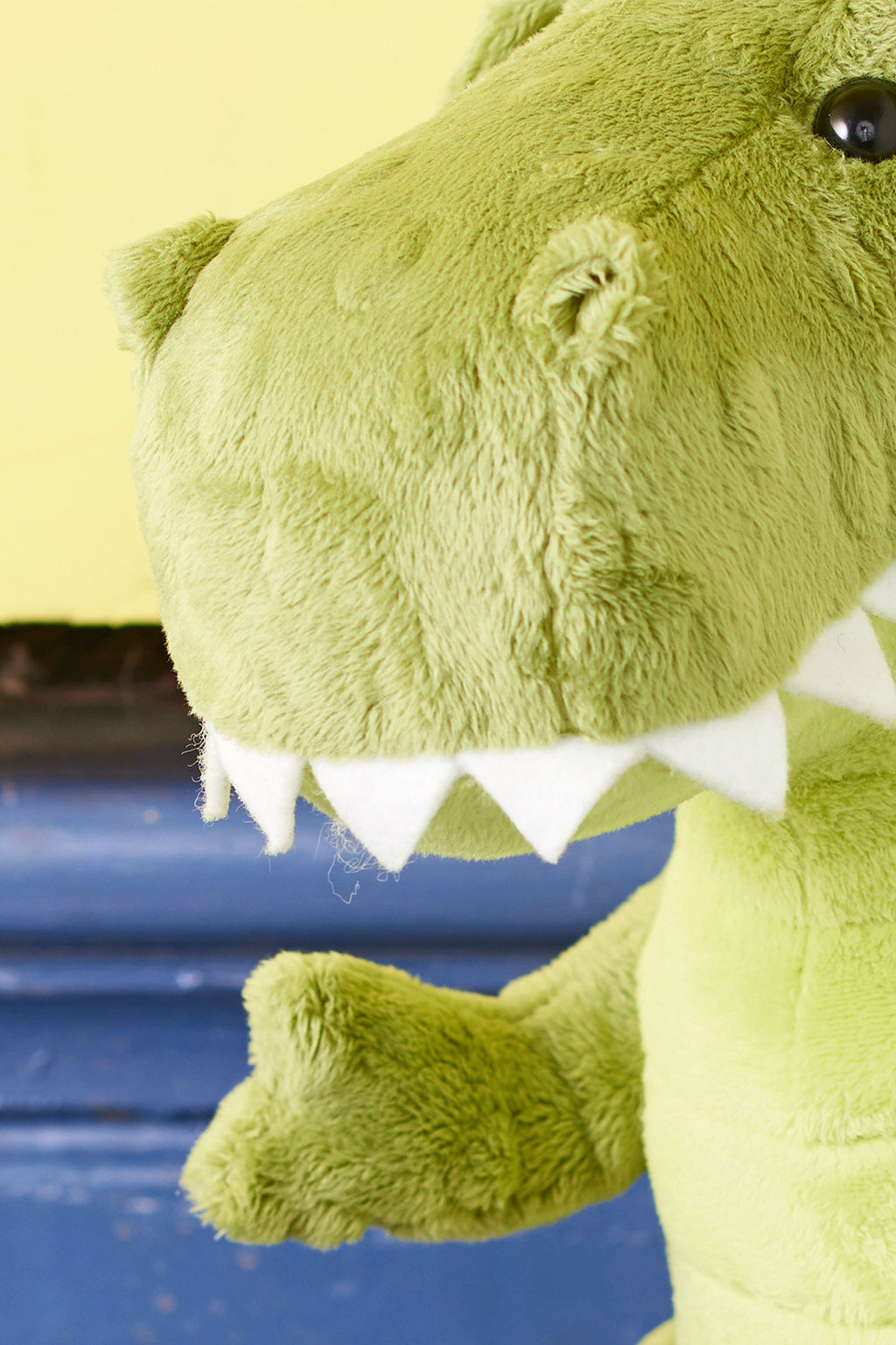 Close-up of a green plush dinosaur toy against a yellow and blue background. The dinosaur's face is partially visible, showcasing its white felt teeth.