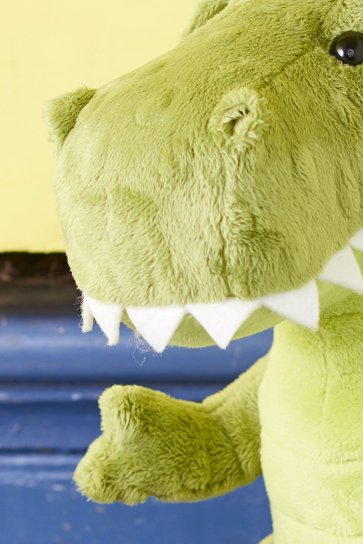 Close-up of a green plush dinosaur toy against a yellow and blue background. The dinosaur's face is partially visible, showcasing its white felt teeth.